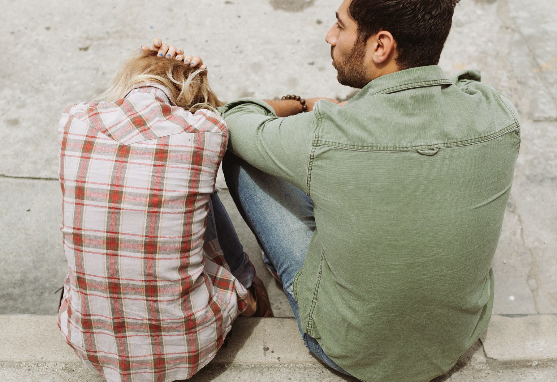 Two people seated against a concrete wall, with one resting their head on their knees.